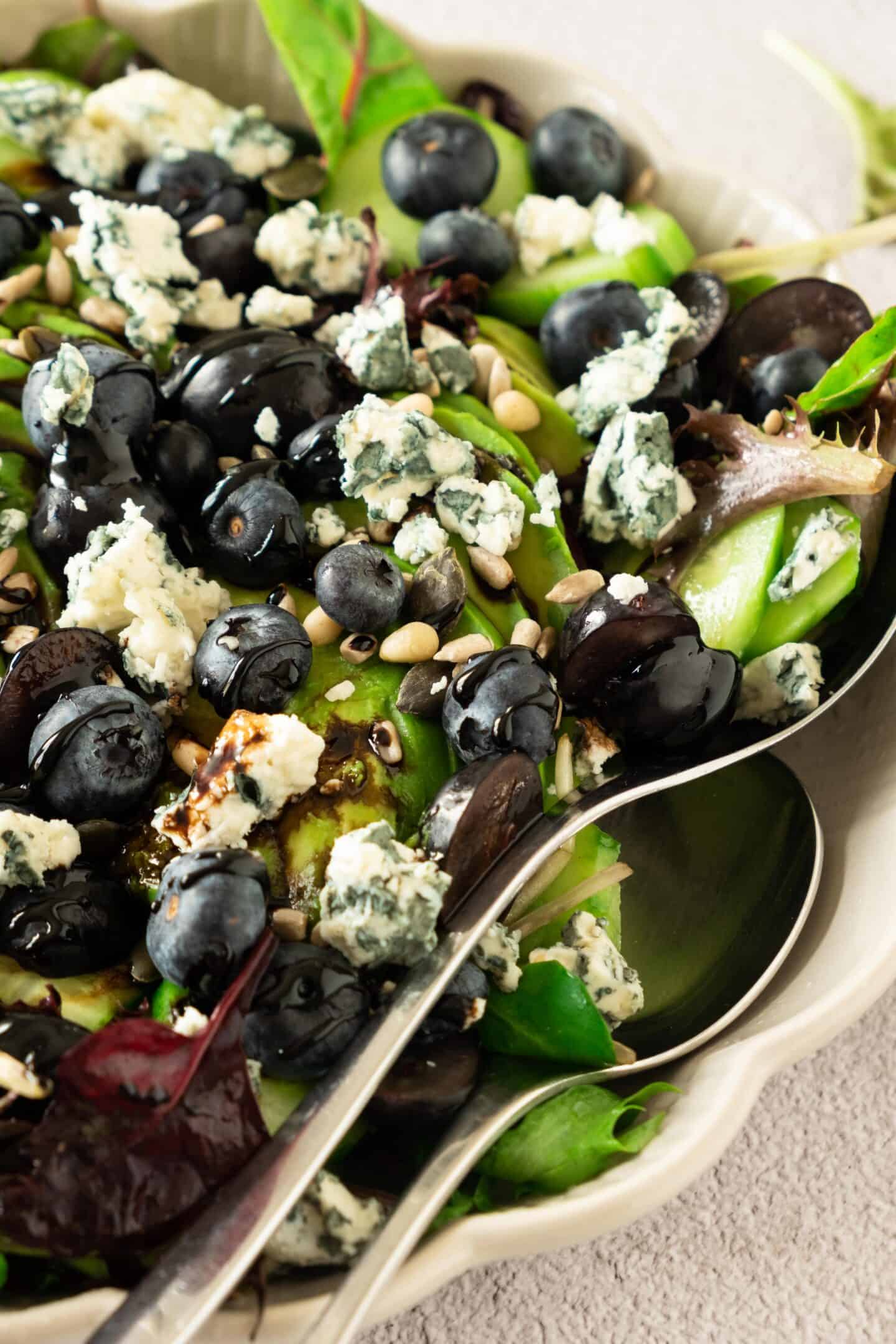 A blueberry salad featuring mixed greens, juicy blueberries, crumbled blue cheese, and pine nuts is served with a metal spoon in a ceramic bowl.