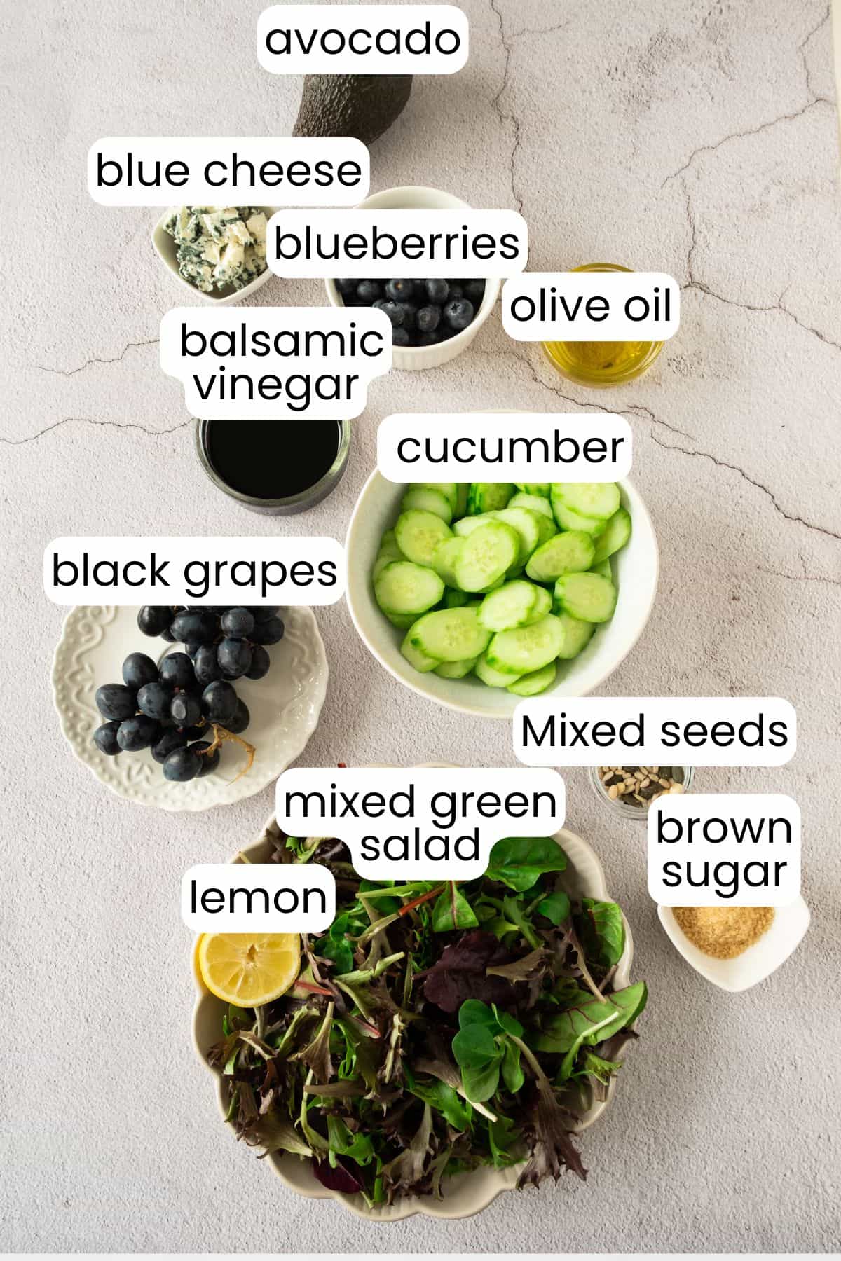 An assortment of fresh salad ingredients, including mixed greens, cucumber, grape and blueberry, creamy avocado, blue cheese, lemon, mixed seeds, olive oil, balsamic vinegar, and a touch of brown sugar.