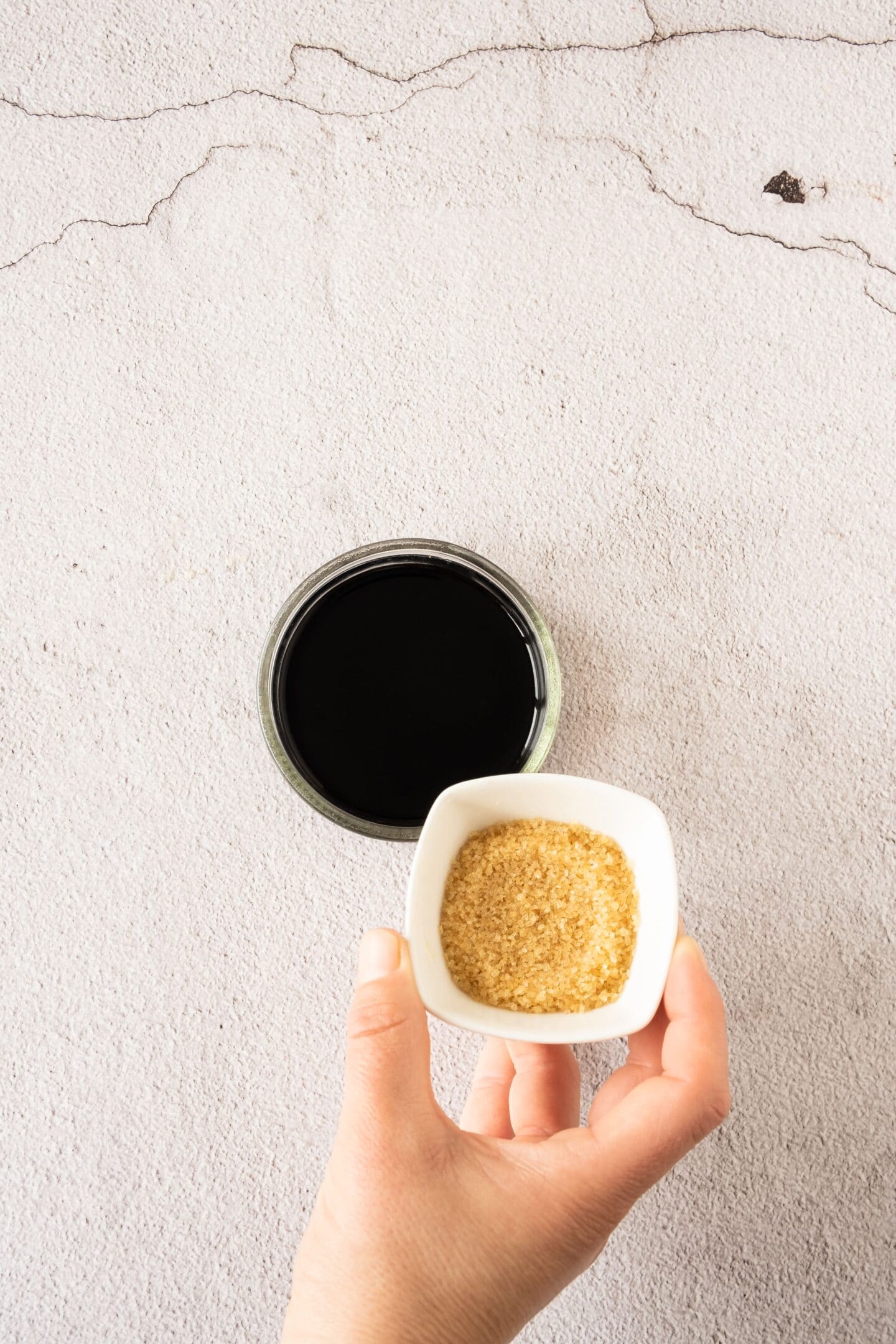 A hand holds a small white bowl of brown sugar above a glass bowl filled with a dark grape liquid on a light textured surface.