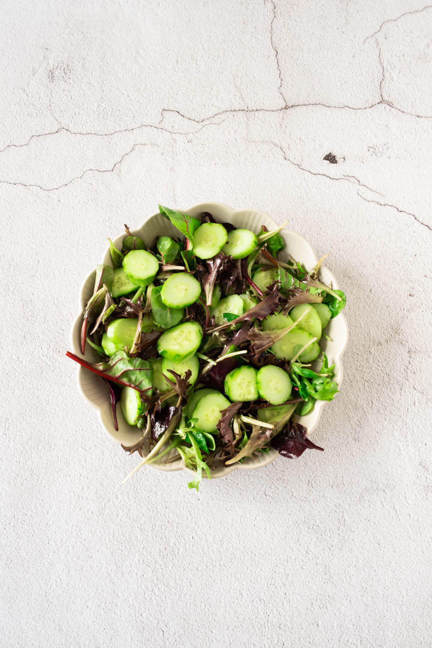 A bowl of mixed green salad with cucumber slices and juicy blueberries on a light, textured surface.