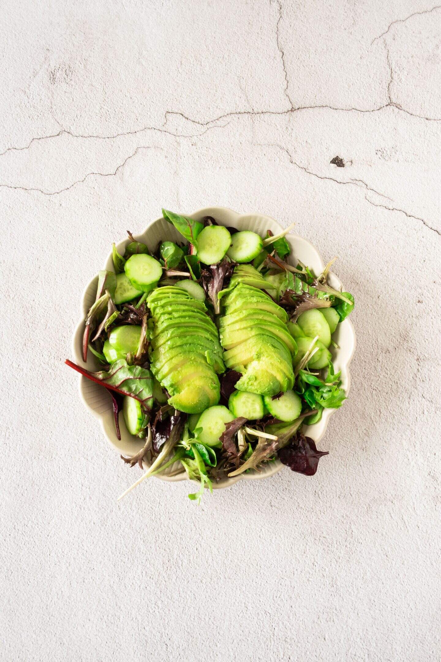 A bowl of mixed green salad topped with sliced avocado, cucumber, and juicy grape halves, served on a light textured surface.