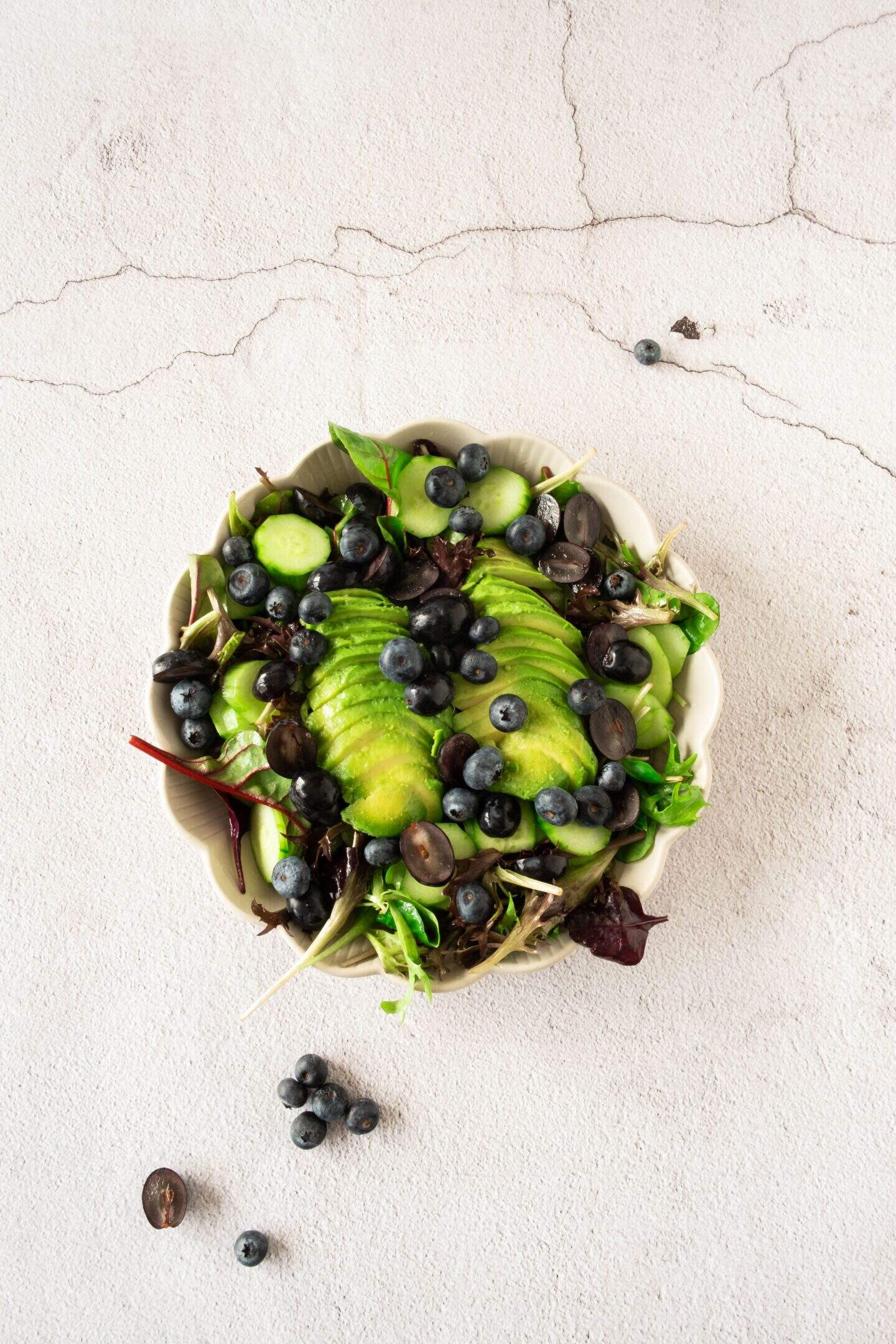 A bowl of salad with sliced avocado, mixed greens, blueberries, and grape halves sits on a light textured surface. Some blueberry and grape pieces are scattered outside the bowl.