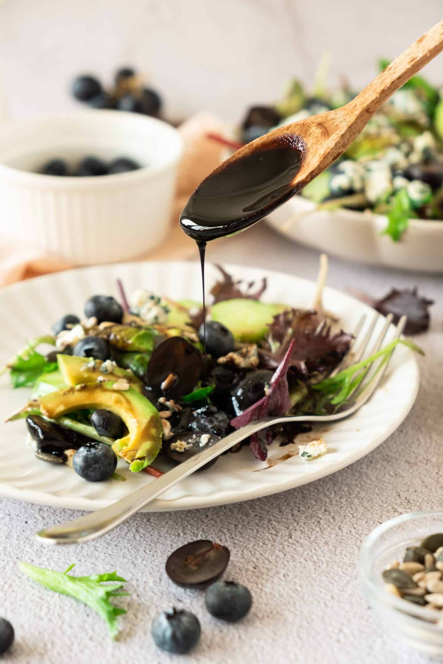 A wooden spoon drizzles dark dressing over a vibrant salad with mixed greens, creamy avocado, juicy grapes, blueberries, and seeds on a white plate.