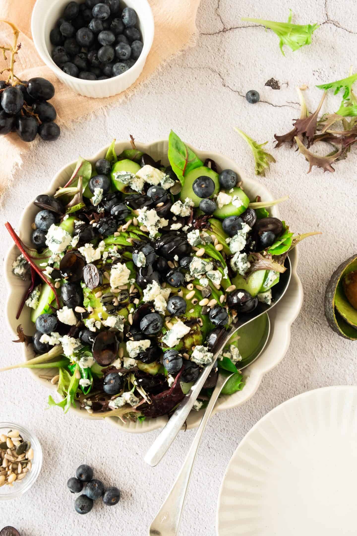 A vibrant salad bowl features mixed greens topped with black grapes, blueberries, crumbled blue cheese, and seeds. A small bowl of fresh blueberries and plates sit nearby, perfect for sharing this colorful grape and blueberry salad.