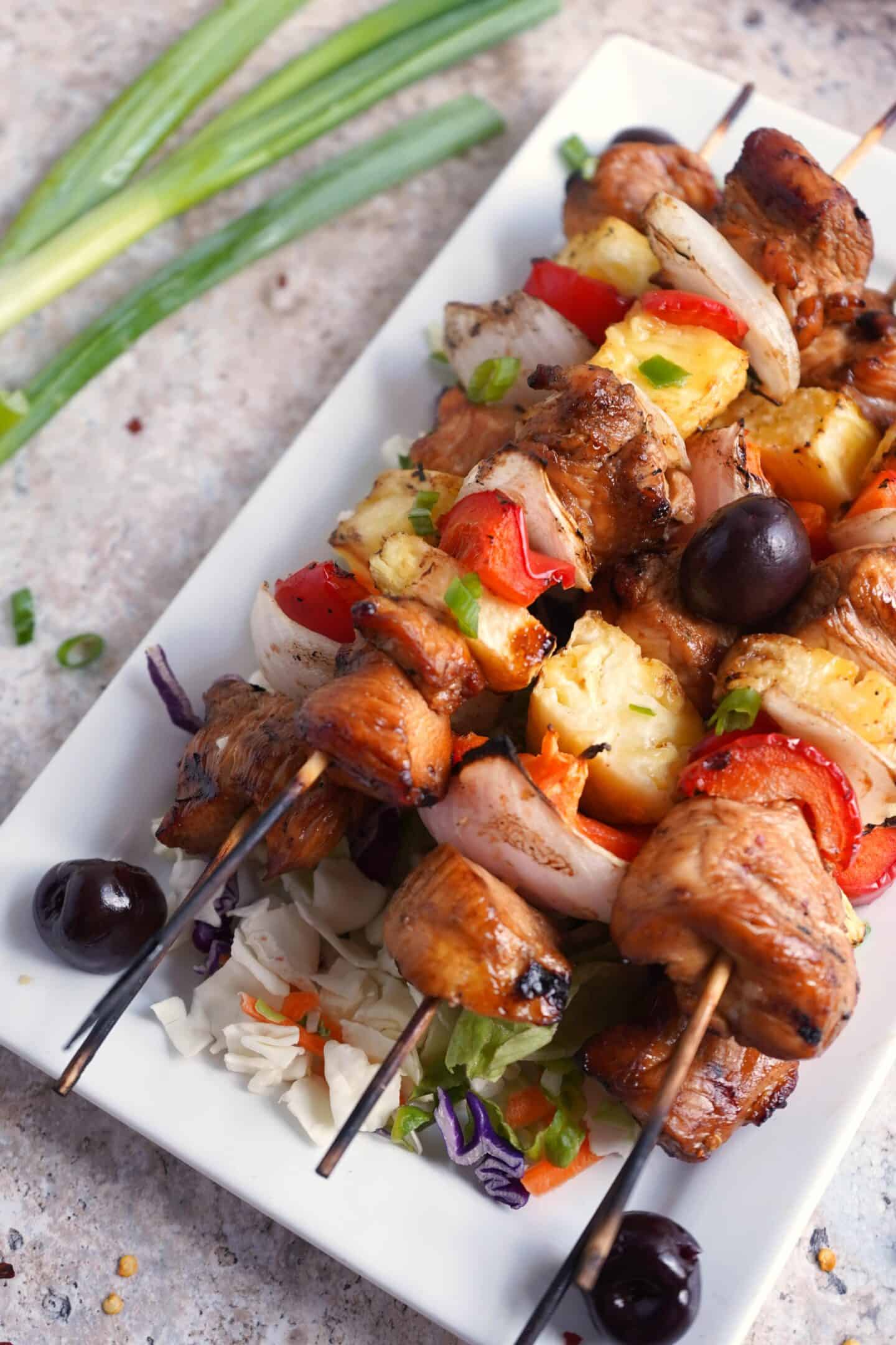 Grilled Chicken Skewers with pineapple, onions, peppers, and olives served on a white plate, accompanied by coleslaw and green onions.