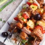 Grilled Chicken Skewers with pineapple, onions, peppers, and olives served on a white plate, accompanied by coleslaw and green onions.