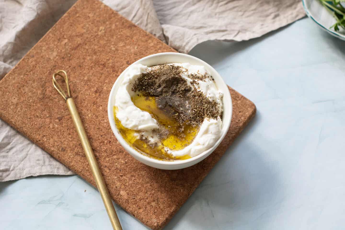 A bowl of labneh made with light yogurt, topped with olive oil, herbs, and spices, sits on a cork board next to a gold spoon and a beige cloth.