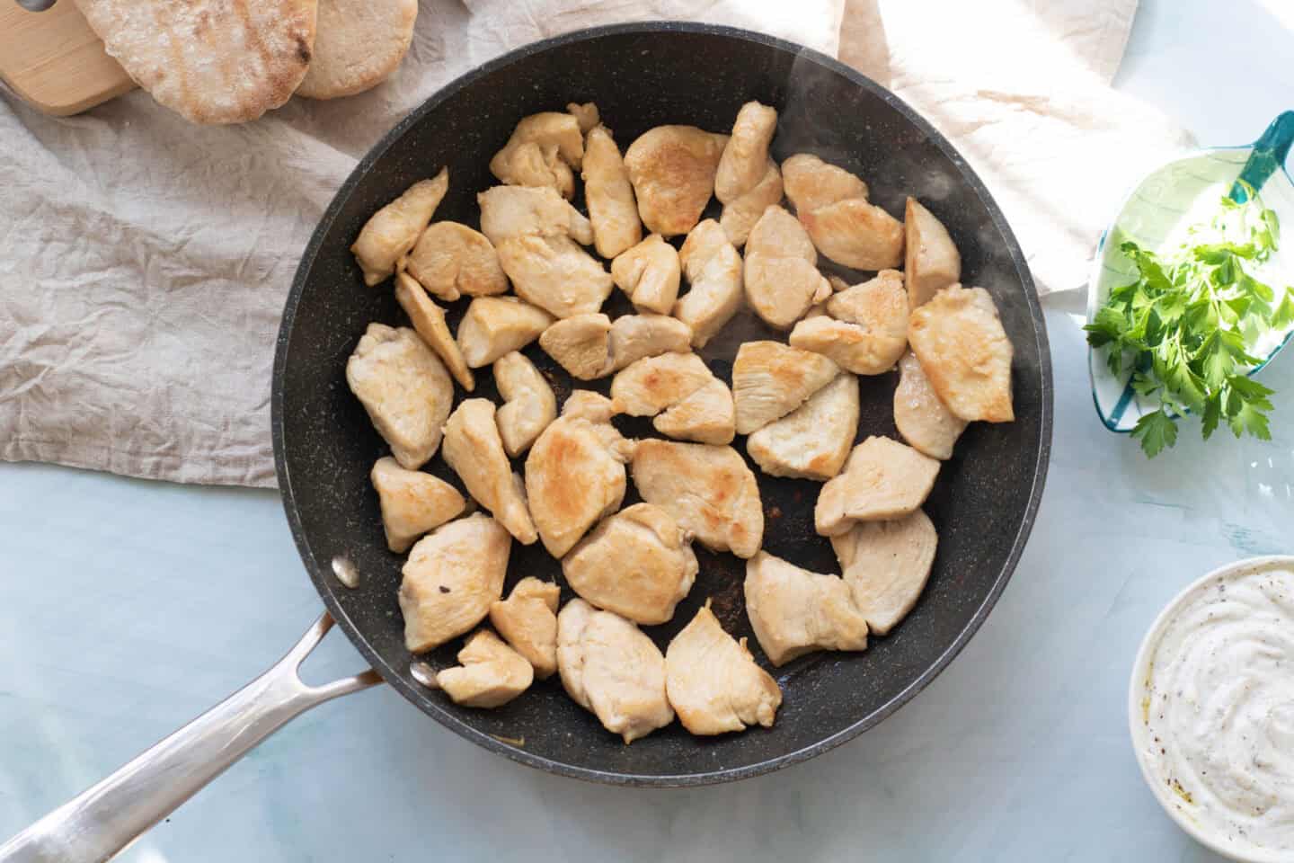 A frying pan filled with cooked, cubed pieces of chicken breast on a light-colored surface, perfect for assembling chicken pitas with bread and herbs nearby—serve with a refreshing light yogurt sauce.