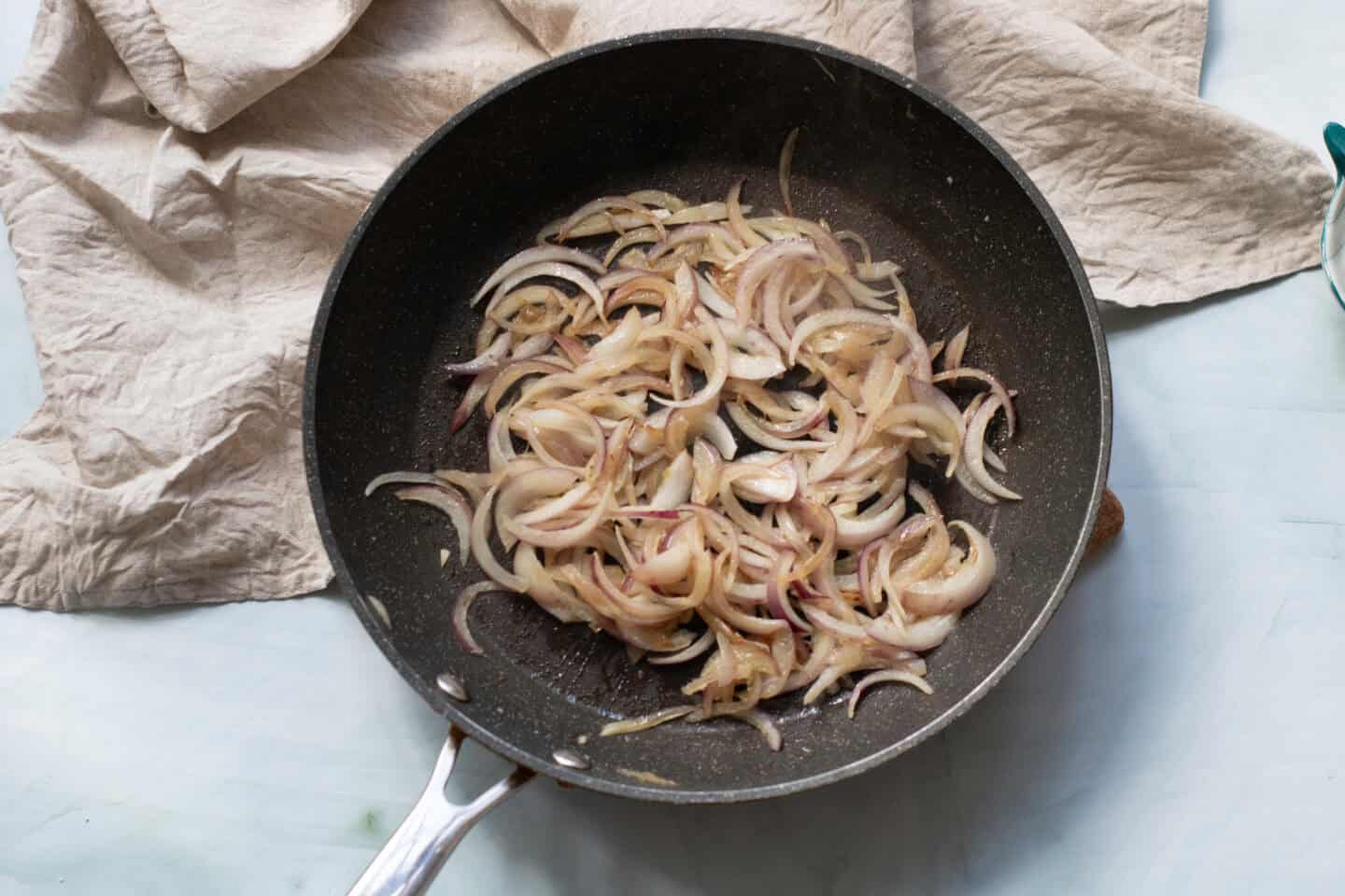 Sliced onions are being sautéed in a black frying pan on a light countertop with a beige cloth nearby, perfect as a base for delicious chicken pitas topped with a refreshing light yogurt sauce.