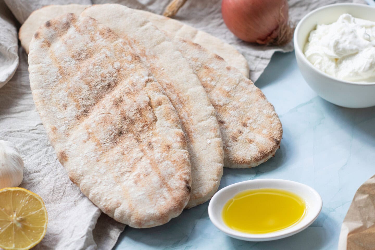 Three pieces of flatbread on a cloth, with a small dish of olive oil, a bowl of light yogurt sauce, a lemon slice, a garlic bulb, and a shallot nearby—perfect accompaniments for chicken pitas.