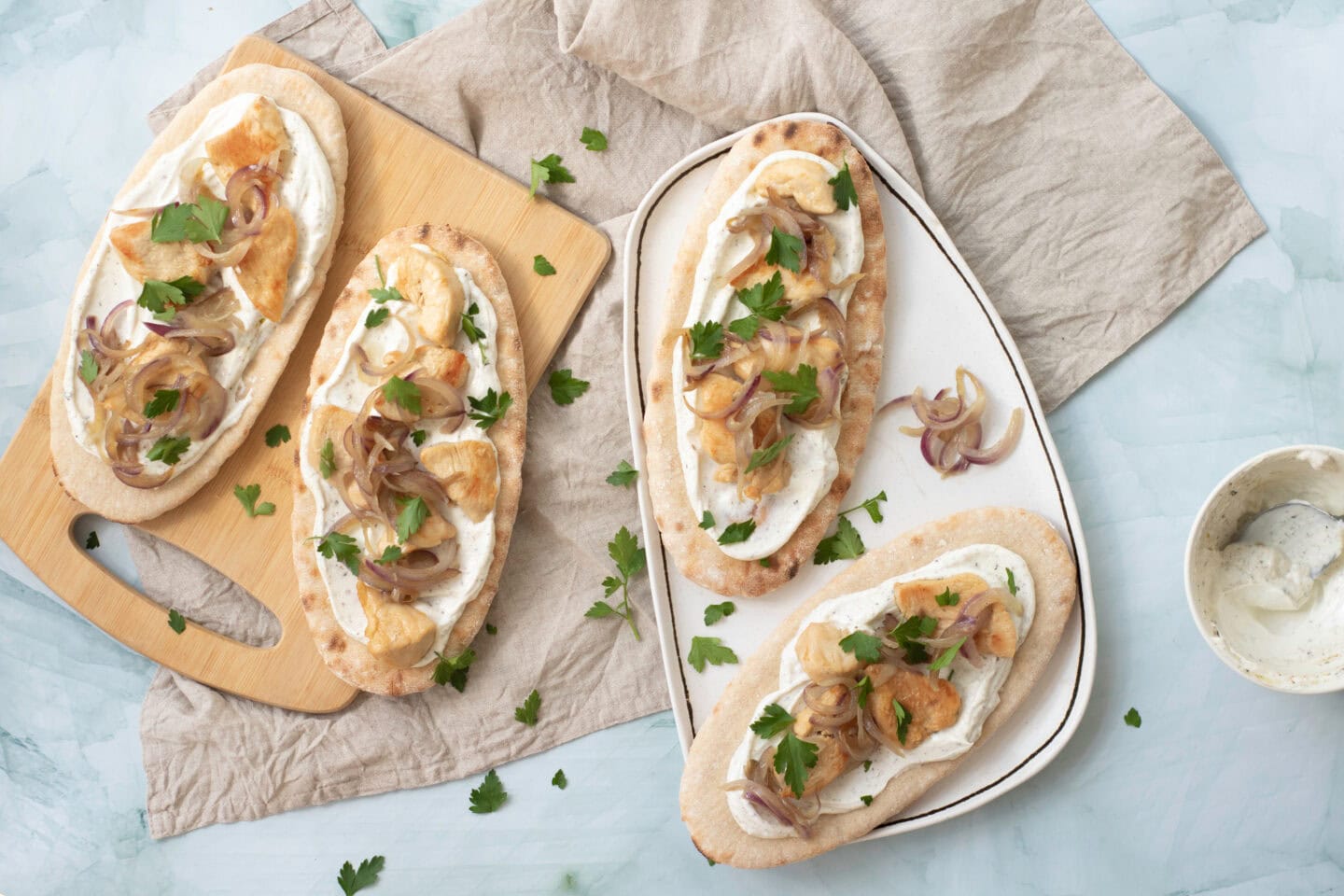 Four flatbreads topped with light sauce, caramelized onions, potato slices, and parsley are arranged on a wooden board and a plate, with a napkin and a small bowl of yogurt sauce nearby.