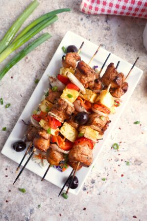 Grilled Chicken Skewers with pineapple, tomatoes, onions, and black olives are served on a white rectangular plate, garnished with green onions nearby.