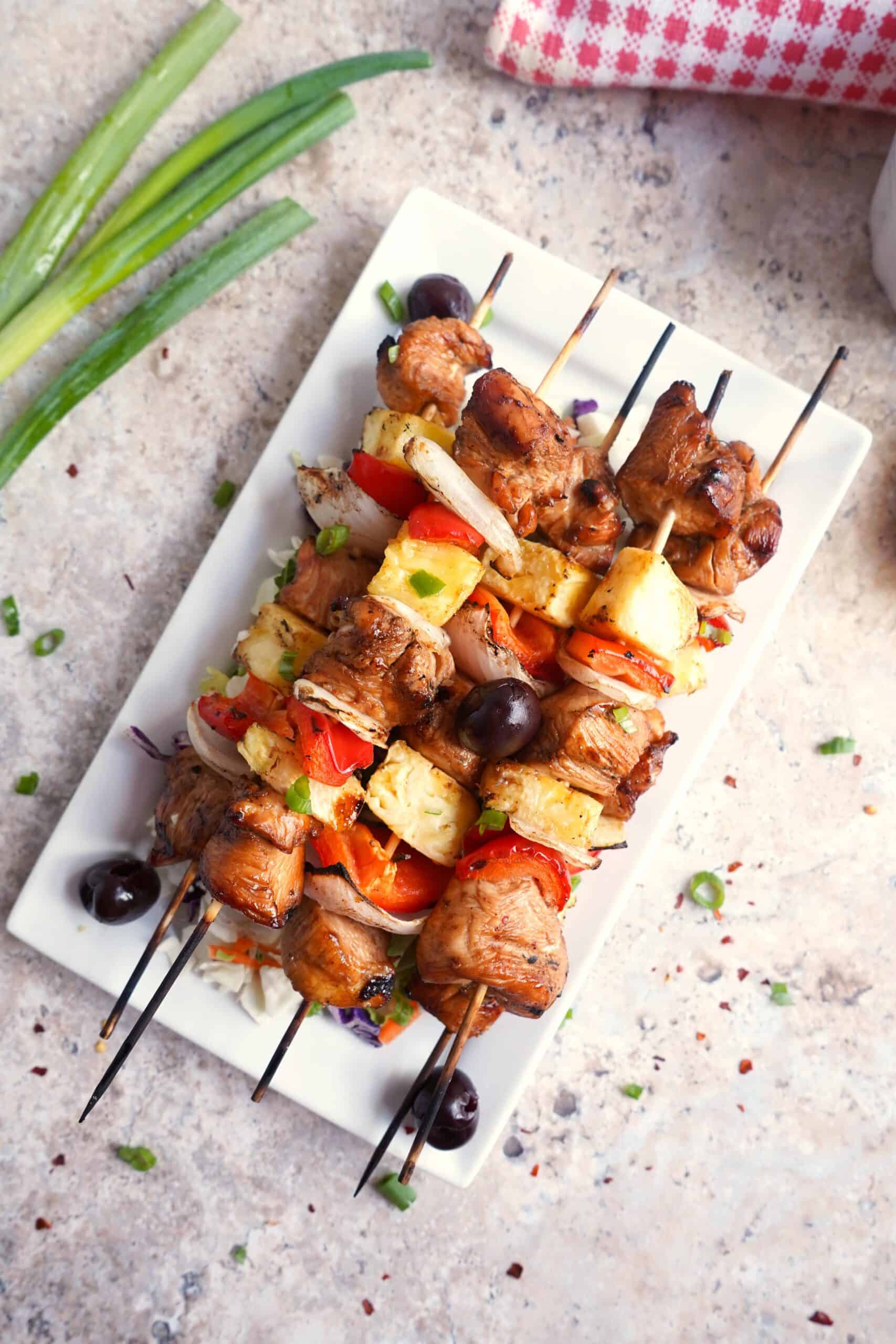 Grilled Chicken Skewers with pineapple, tomatoes, onions, and black olives are served on a white rectangular plate, garnished with green onions nearby.