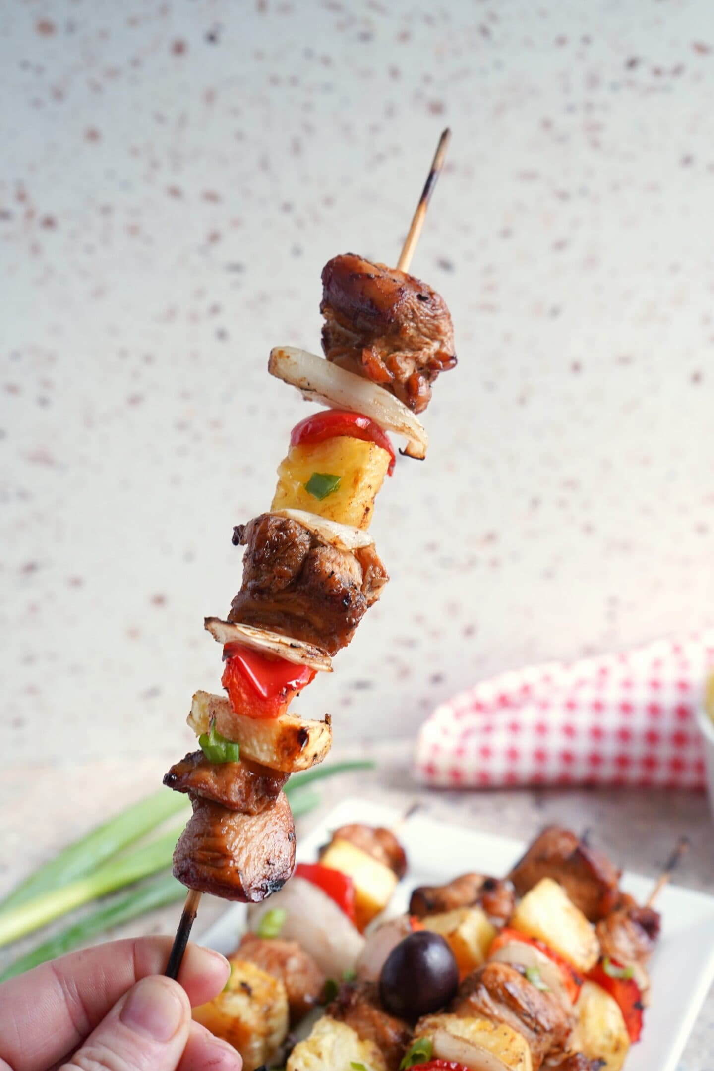A hand holds a skewer with grilled chicken, pineapple, onions, and peppers; more grilled chicken skewers are visible on a plate in the background.