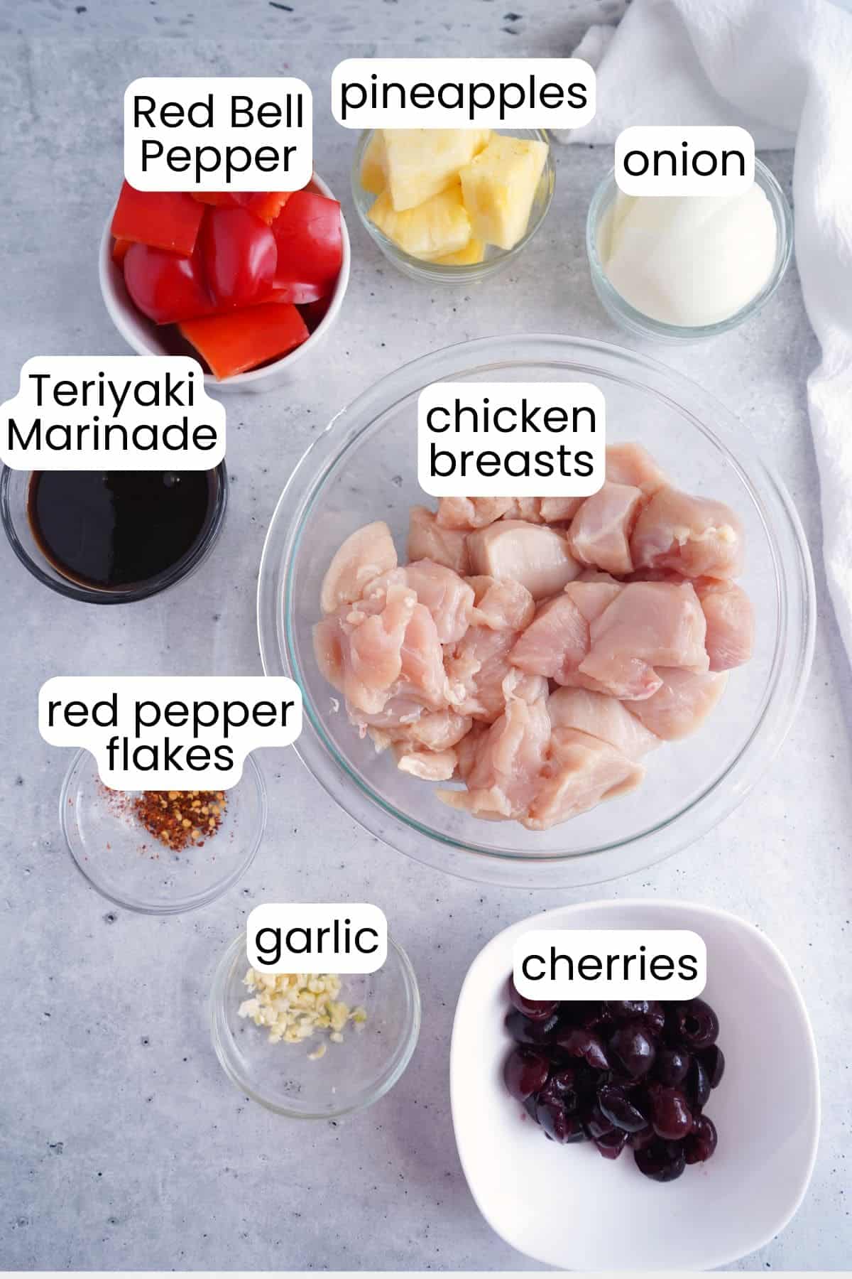 Bowls containing ingredients for Cherry Chicken Teriyaki—raw chicken breasts, red bell pepper, pineapple, onion, teriyaki marinade, red pepper flakes, garlic, and cherries—are arranged on a gray surface.