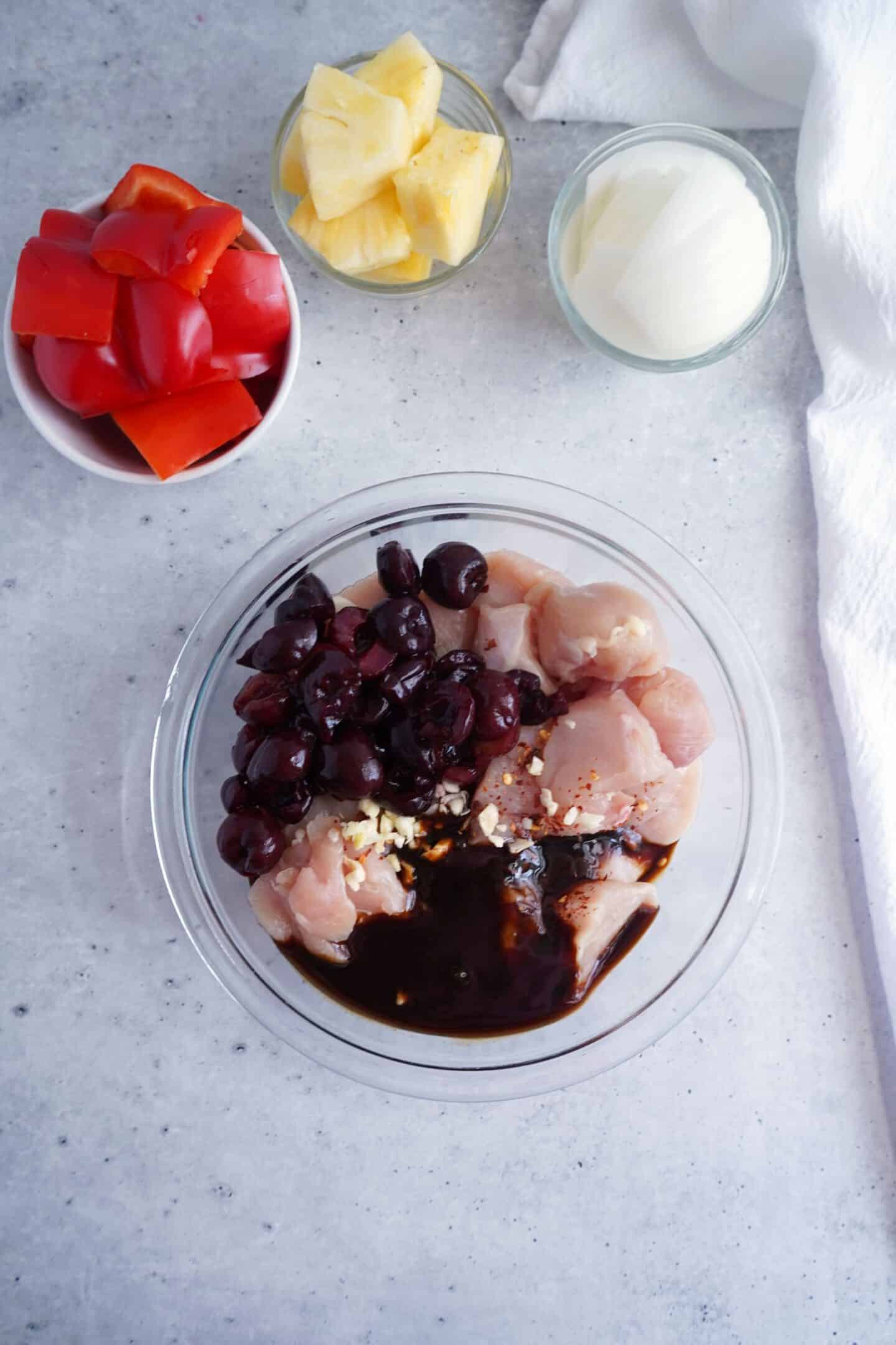 A glass bowl of raw Cherry Chicken with cherries, garlic, and dark sauce, alongside small bowls of red bell pepper, pineapple, and onion on a light gray surface.