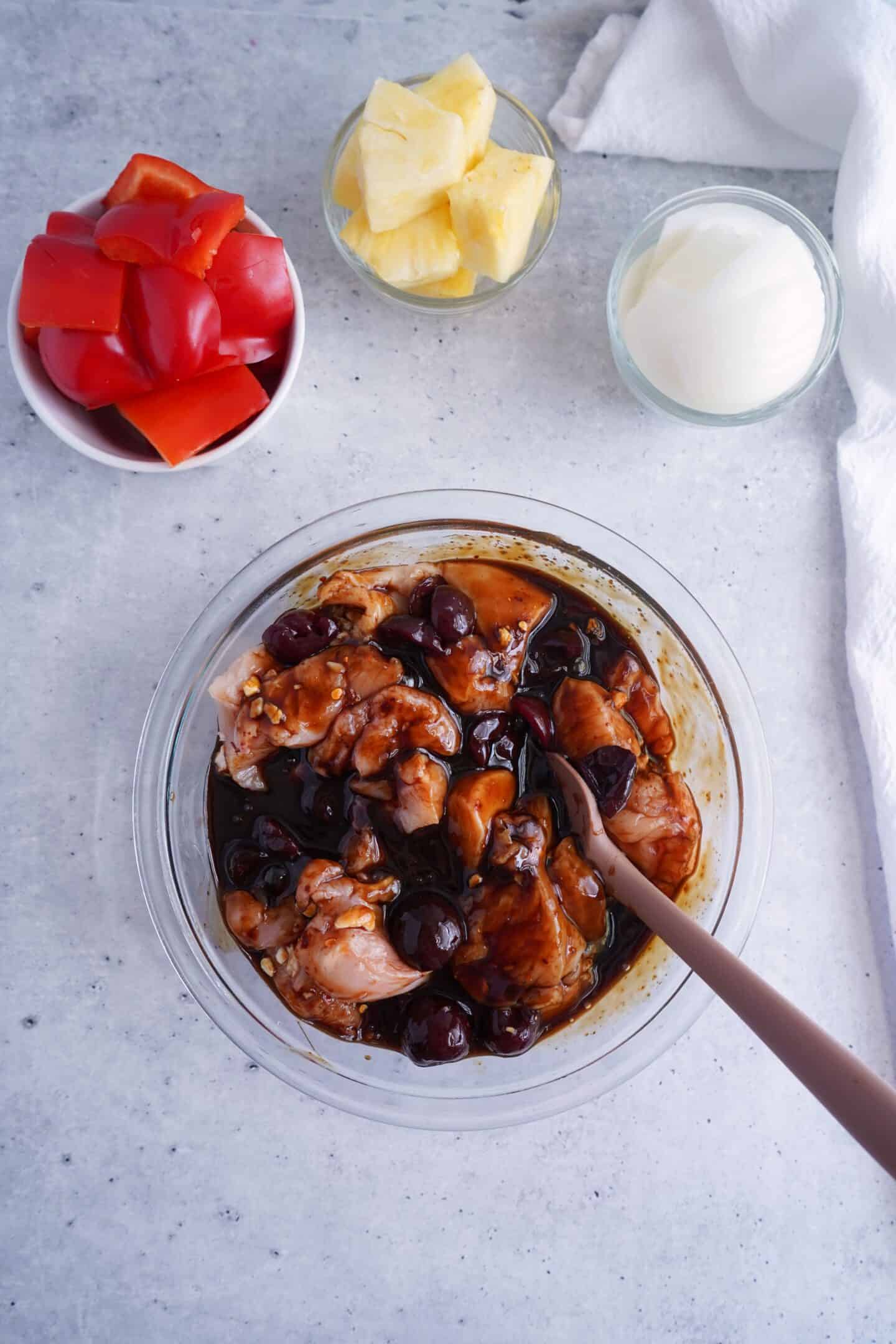 A bowl of raw Cherry Chicken pieces marinating in a dark sauce with cherries, surrounded by small bowls of red bell pepper, pineapple chunks, and onion.