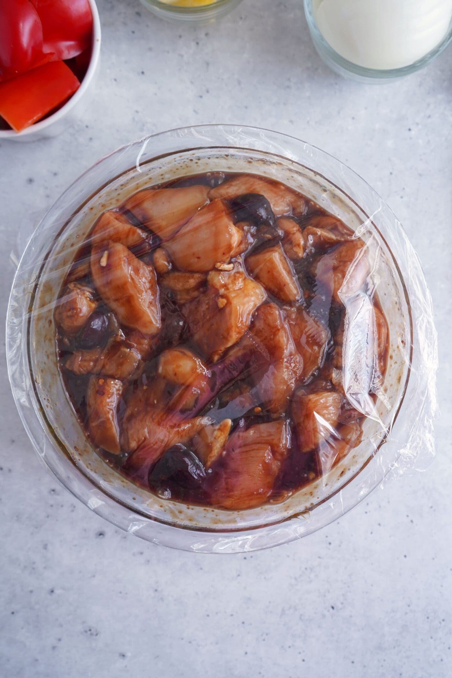 A glass bowl of marinating Teriyaki Chicken pieces covered with plastic wrap sits on a light countertop, with red bell peppers and an onion nearby—perfect prep for flavorful Grilled Chicken Skewers.