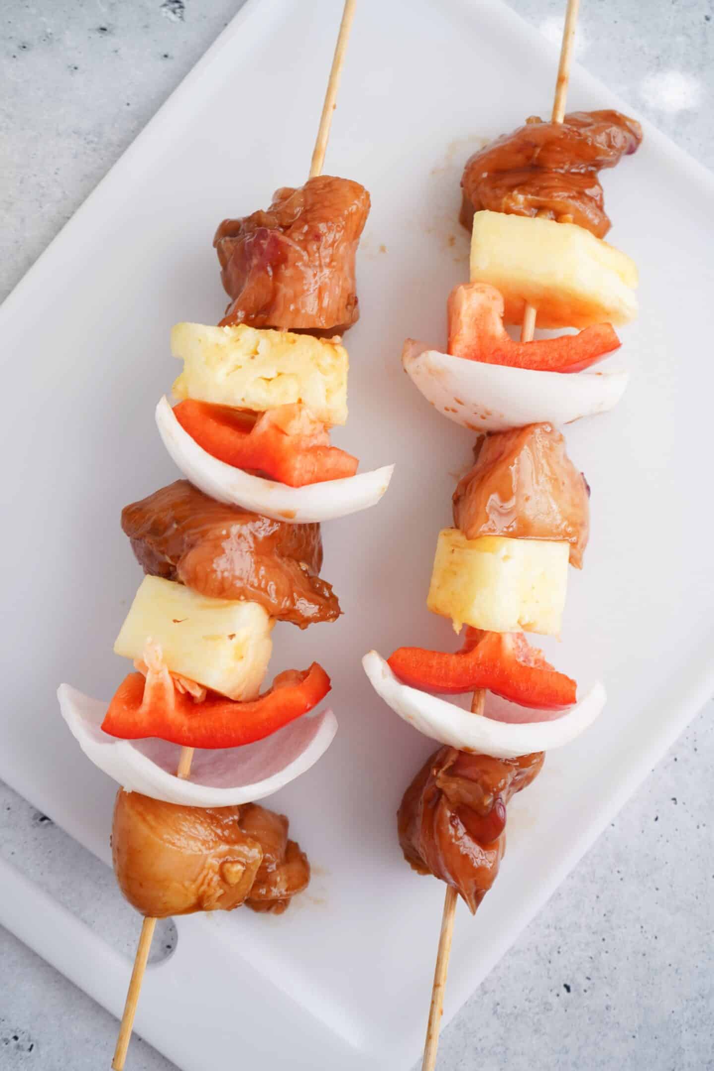 Two skewers with marinated Grilled Chicken Skewers, pineapple chunks, red bell pepper, and onion on a white cutting board.