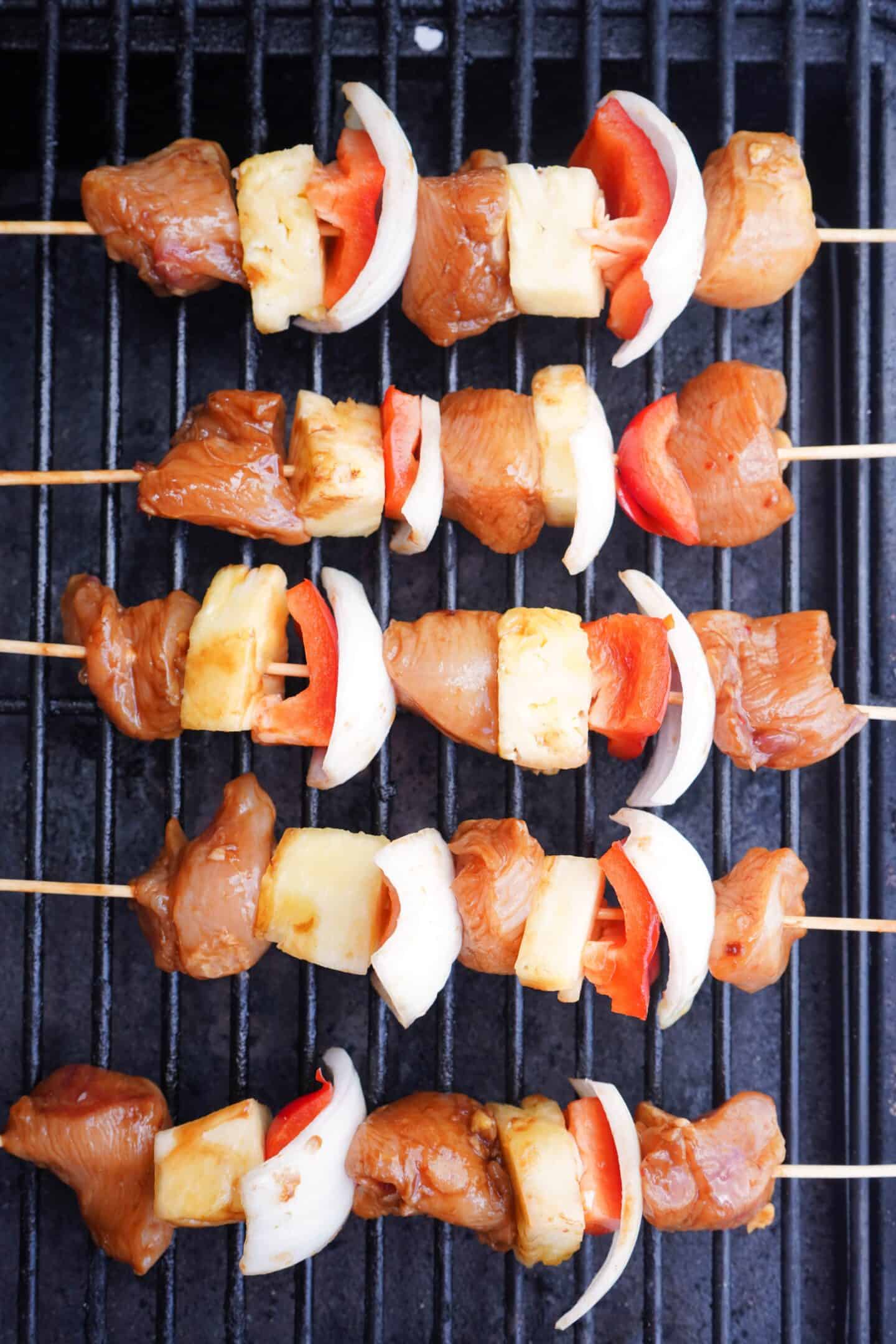 Three skewers with marinated grilled chicken, pineapple, onion, and red bell pepper pieces are placed on a grill, ready to be cooked.
