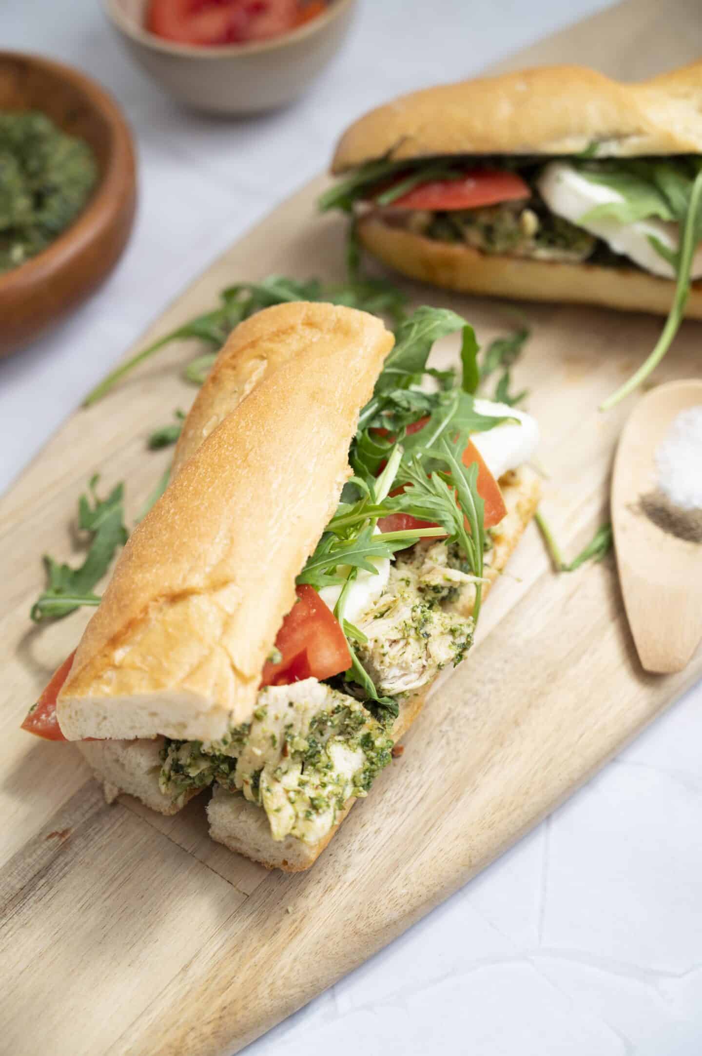 A Chicken Sandwich filled with tomato slices, mozzarella, arugula, and walnut pesto sits on a wooden board next to a small pile of salt.