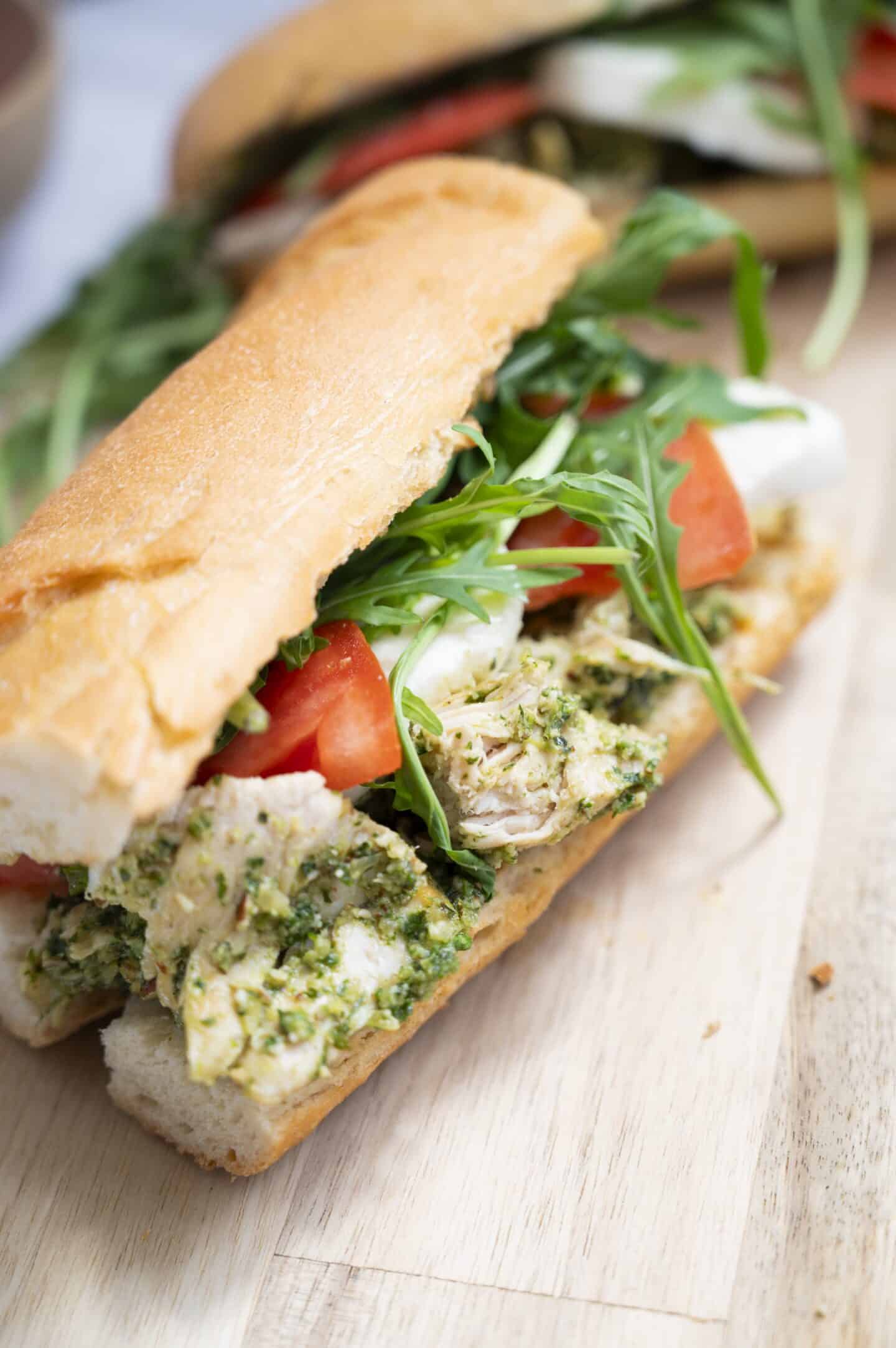 A baguette sandwich filled with shredded chicken in walnut pesto, fresh tomato slices, mozzarella cheese, and arugula, placed on a wooden surface.