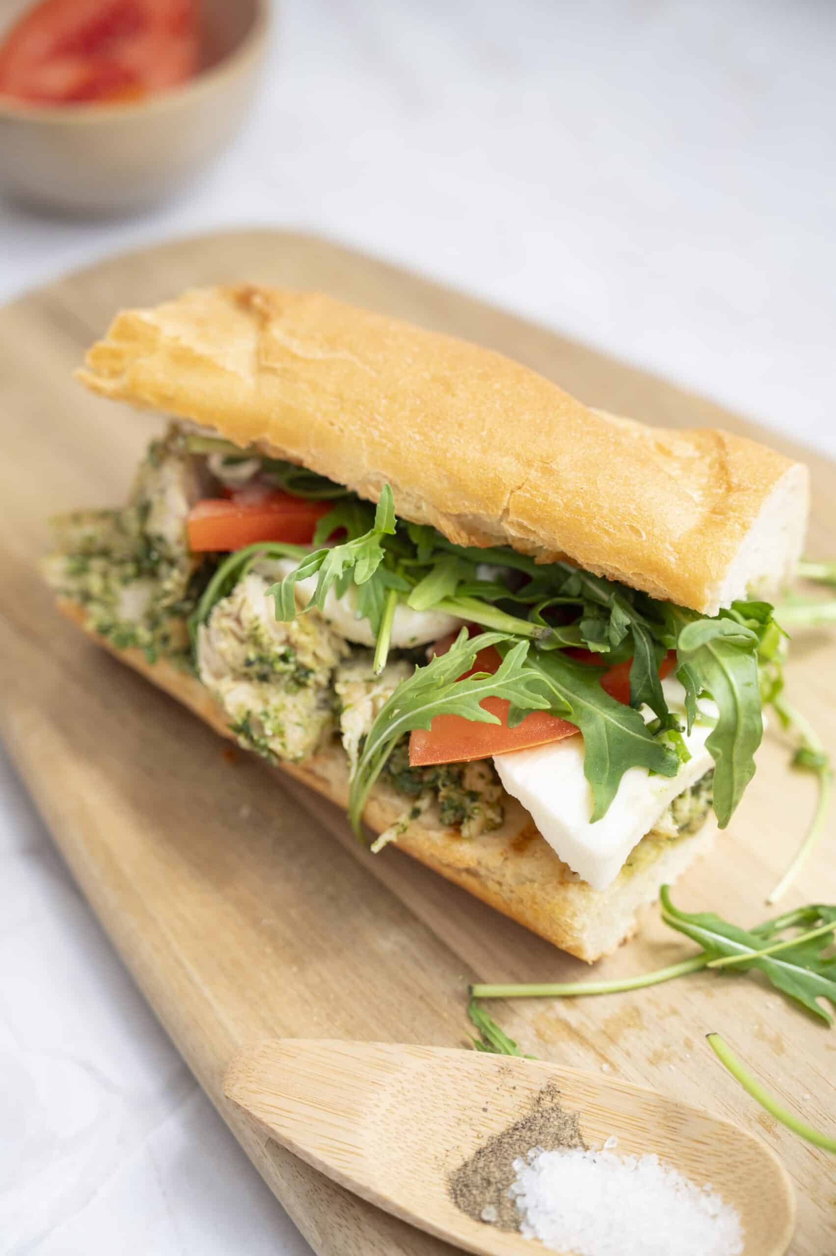 A Chicken Sandwich with mozzarella, tomato slices, arugula, and walnut pesto on a baguette sits on a wooden board, with salt, pepper, and a bowl of tomatoes nearby.