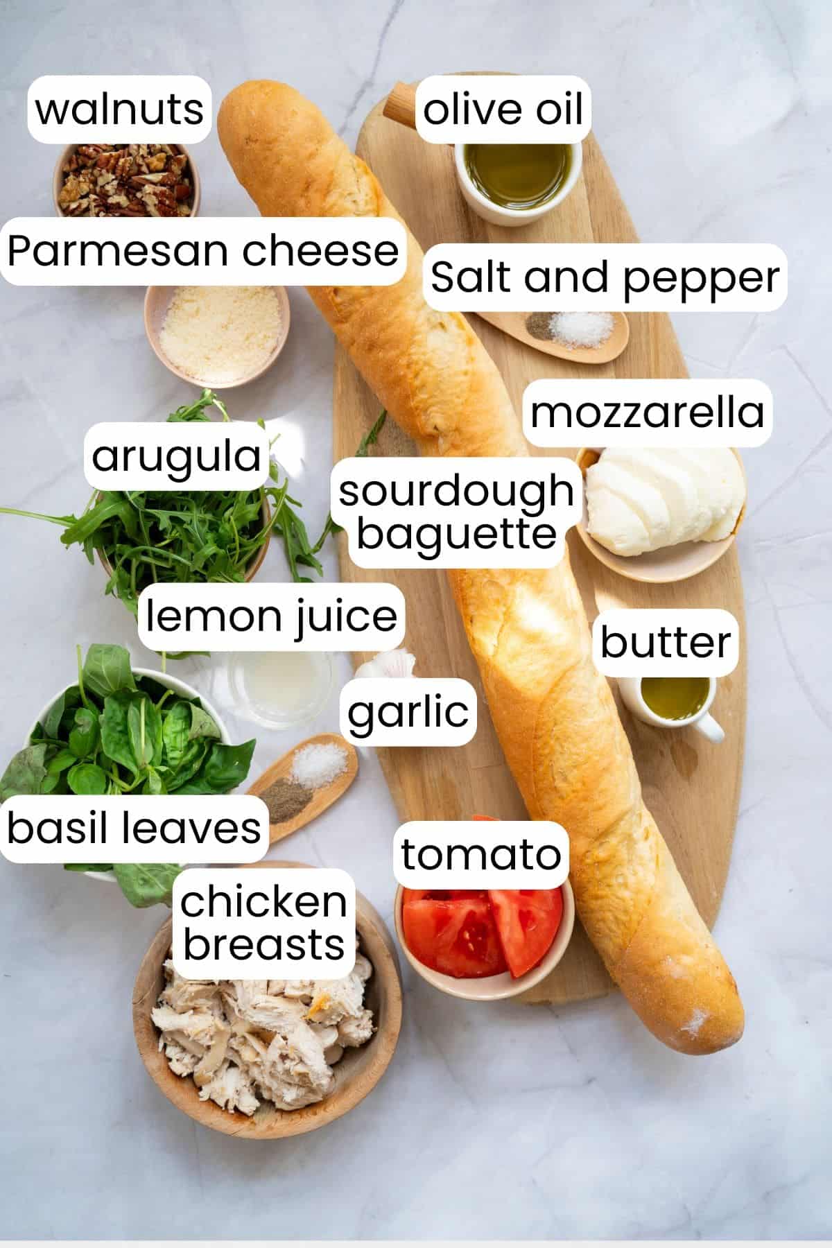 A sourdough baguette on a board surrounded by labeled ingredients: chicken, tomato, basil, arugula, lemon juice, walnuts, Parmesan, mozzarella, olive oil, butter, garlic, salt, and pepper—perfect for a Walnut Pesto Chicken Sandwich.