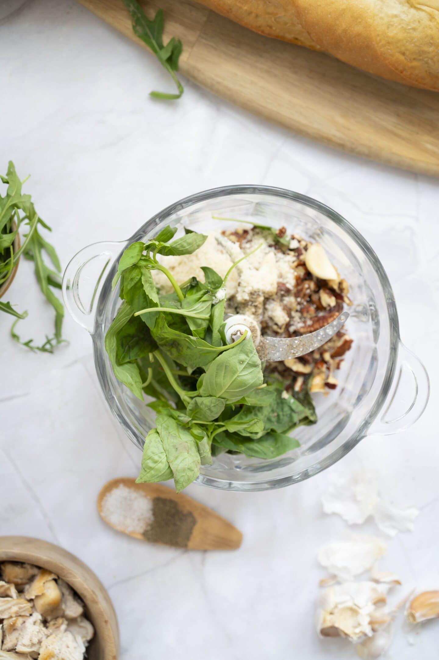 A glass food processor bowl filled with basil leaves, nuts, grated cheese, and seasonings sits on a light countertop near a loaf of bread, ready to create a flavorful walnut pesto for the perfect chicken sandwich.