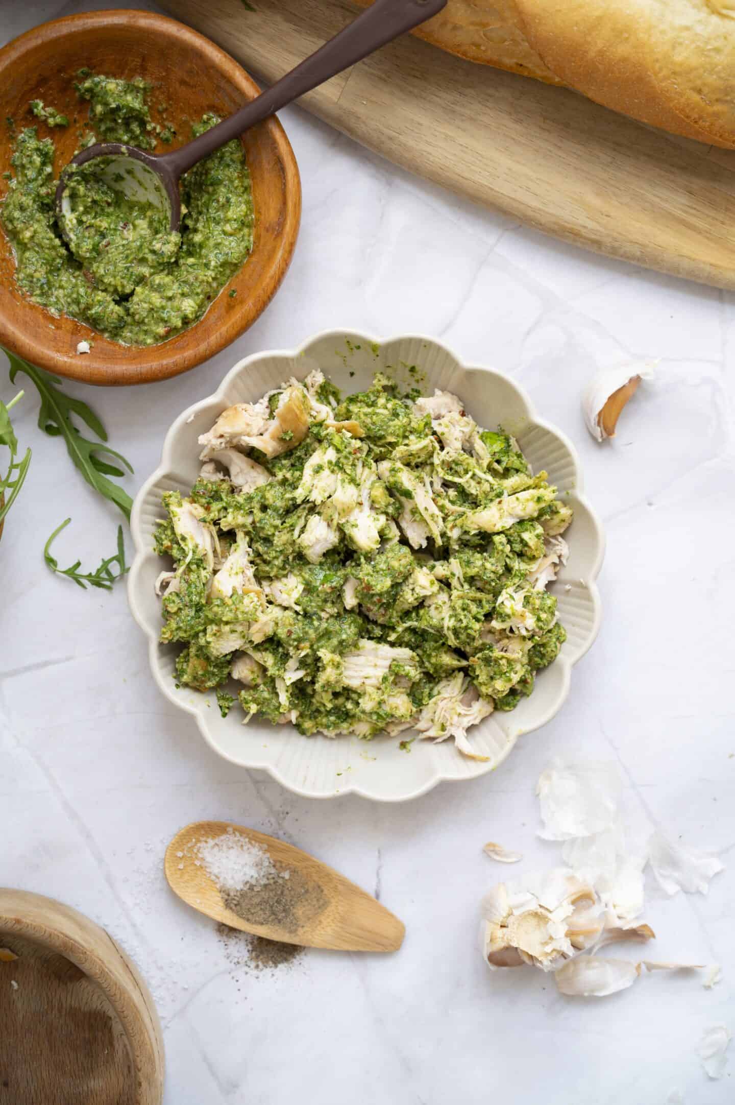 Shredded chicken mixed with green pesto in a scalloped bowl, perfect for a Chicken Walnut Pesto Sandwich, surrounded by a wooden bowl of pesto, garlic cloves, ground pepper, and a loaf of bread on a marble surface.