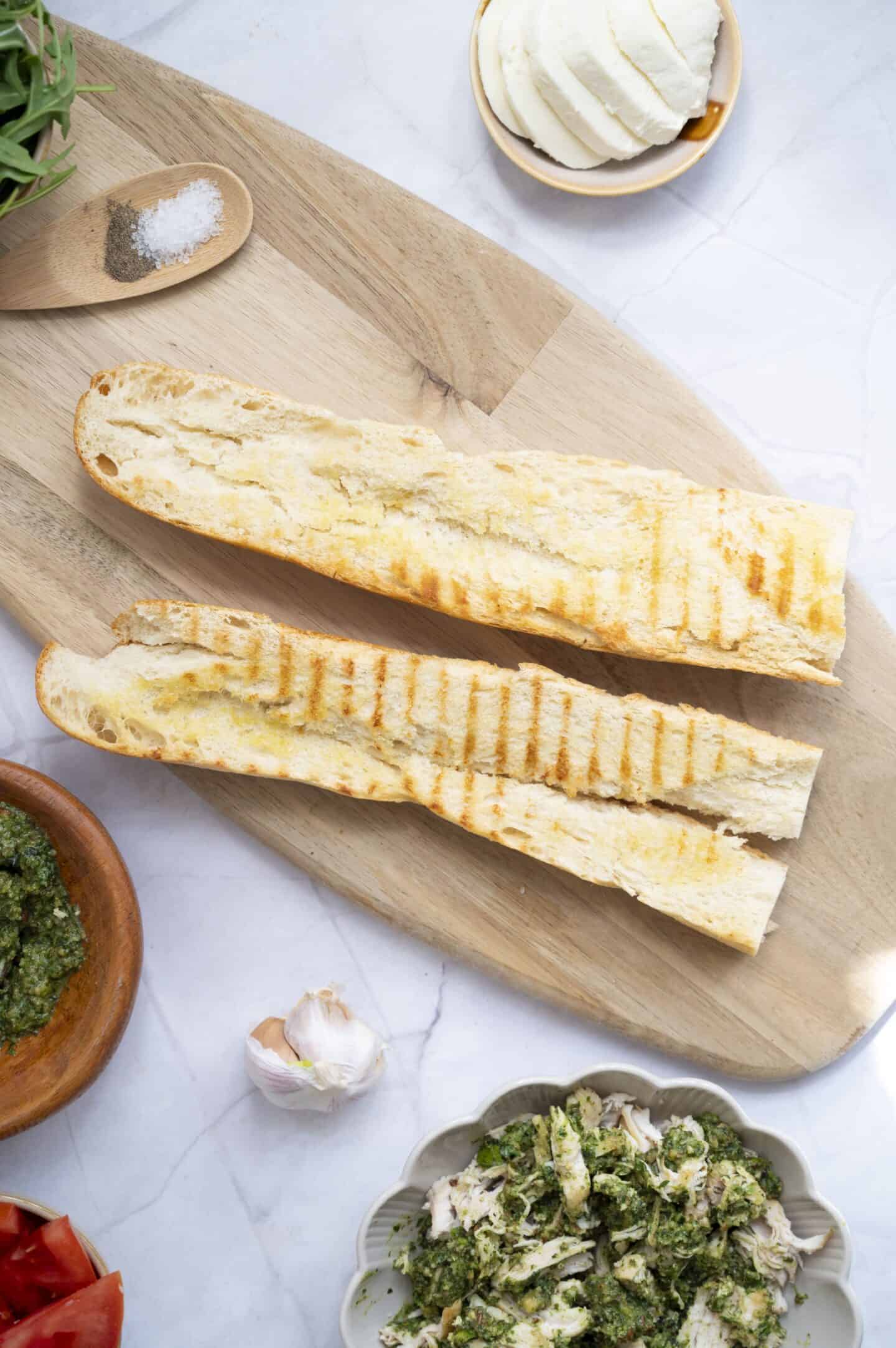 A sliced, grilled baguette on a wooden board surrounded by mozzarella, tomato slices, garlic, shredded chicken with walnut pesto, and a wooden spoon with salt—perfect for crafting a flavorful chicken sandwich.