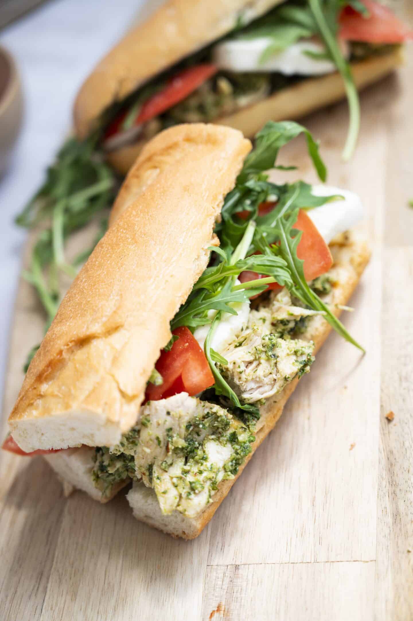 A Chicken Walnut Pesto Sandwich on a baguette, filled with chicken, tomatoes, mozzarella cheese, pesto, and fresh arugula sits on a wooden surface.