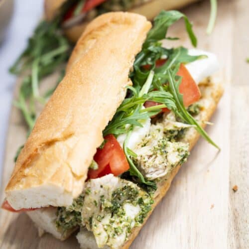 A Chicken Walnut Pesto Sandwich on a baguette, filled with chicken, tomatoes, mozzarella cheese, pesto, and fresh arugula sits on a wooden surface.