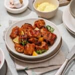 A plate of grilled marinated pork skewers garnished with green onions and lemon wedges, surrounded by bowls of homemade aioli, sauce, sliced chilies, and lemon quarters on a table.