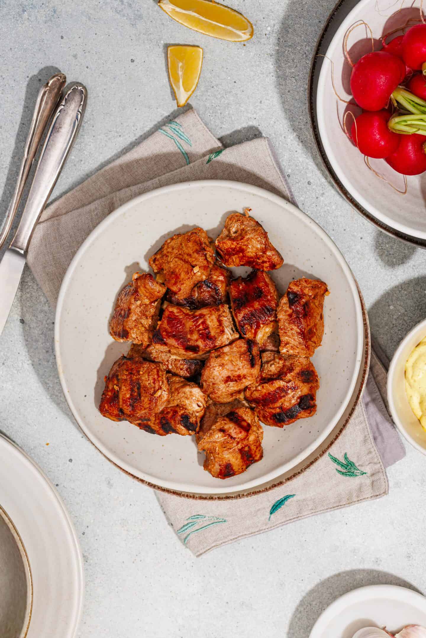 A plate of Grilled Marinated Pork Skewers rests on a beige napkin, served with lemon wedges, radishes, and a bowl of Homemade Aioli on a light surface.