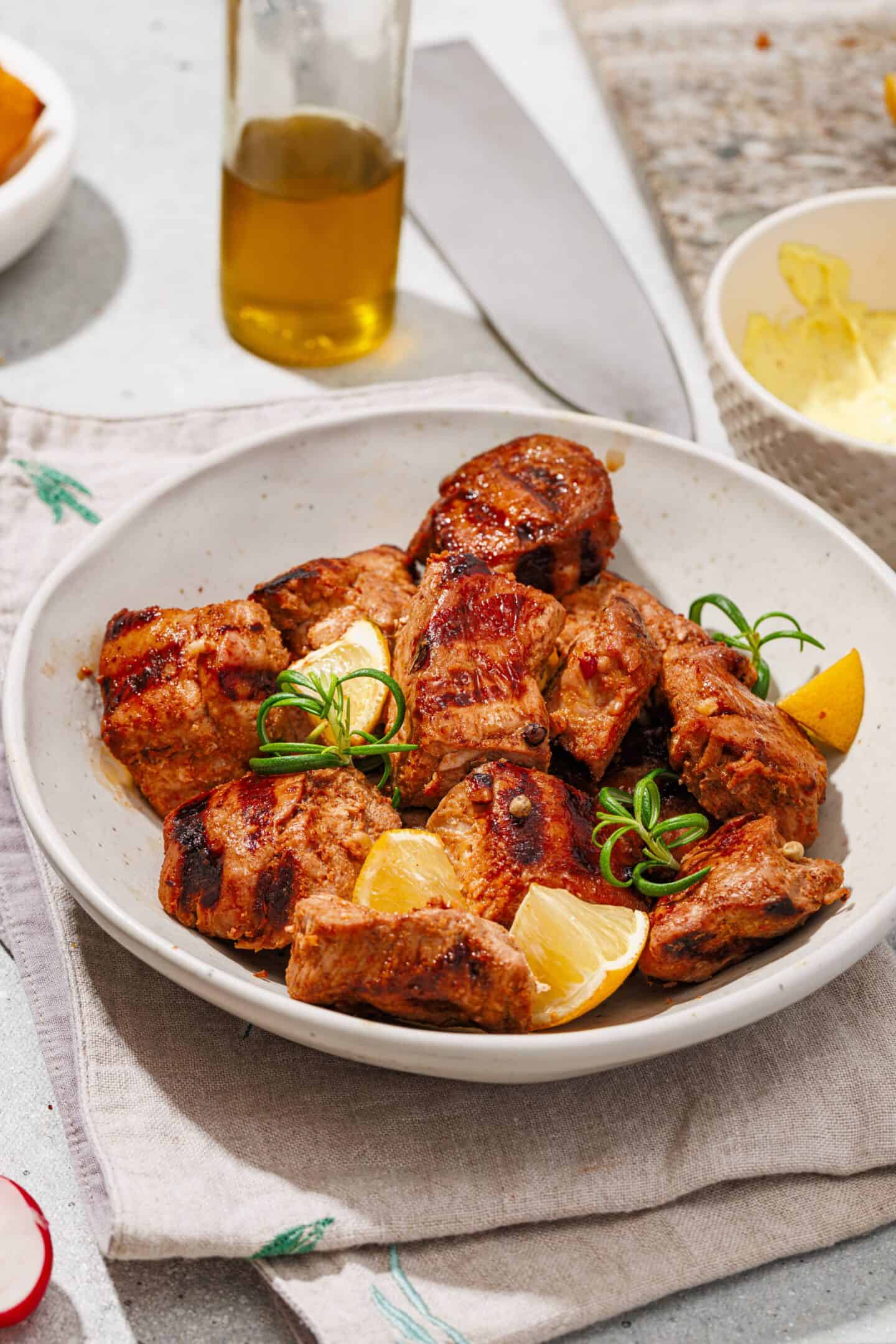 A white bowl containing grilled pork skewers of seasoned meat, garnished with lemon wedges and herbs, placed on a napkin with homemade aioli, a bottle of oil, and a knife in the background.