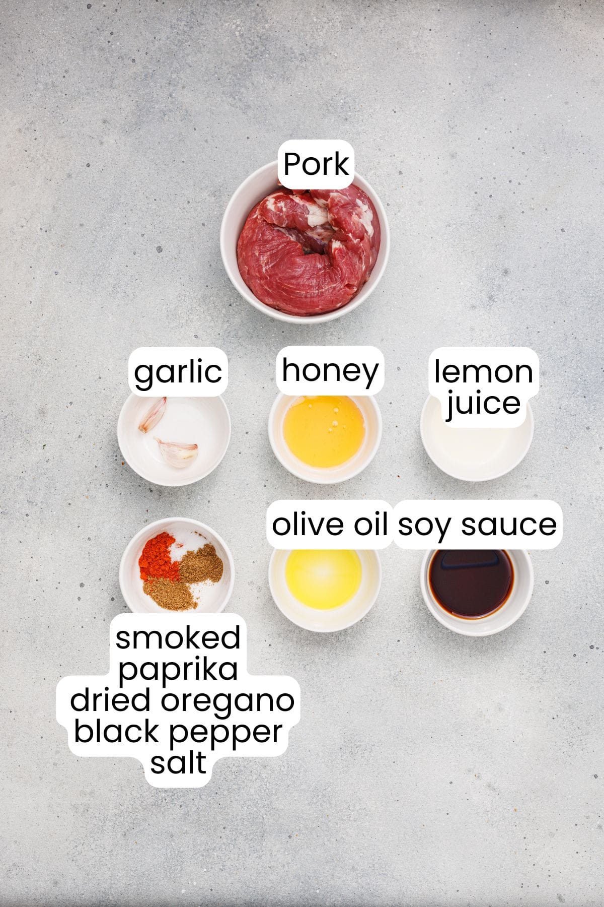 Top-down view of individual bowls with ingredients for Marinated Pork: pork, garlic, honey, lemon juice, olive oil, soy sauce, and spices on a gray surface—perfect for Grilled Pork Skewers with Homemade Aioli.