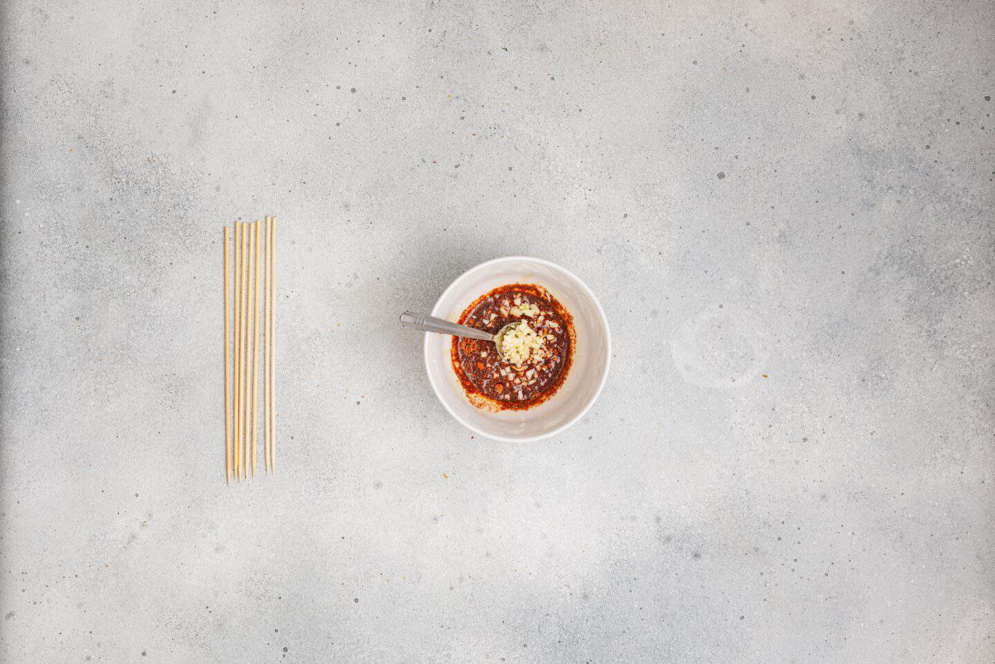 A bowl of spicy sauce with a spoon, garnished with minced garlic and sesame seeds, sits next to uncooked noodles—perfect for pairing with Grilled Marinated Pork Skewers with Homemade Aioli on a light gray surface.