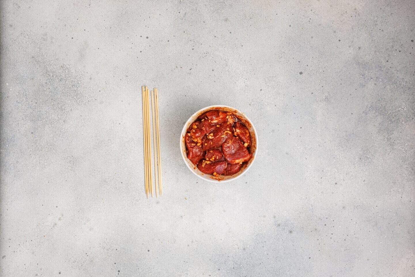 A bowl of Grilled Marinated Pork Skewers with Homemade Aioli sits next to four uncooked spaghetti noodles on a light gray textured surface.