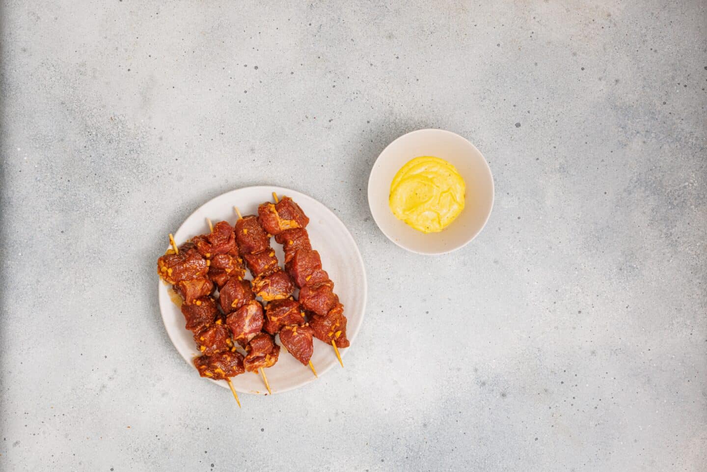 A plate of grilled marinated pork skewers is placed next to a bowl of homemade aioli on a light gray surface.