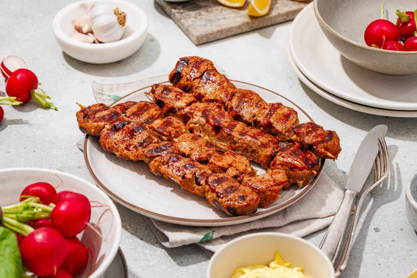 Grilled Marinated Pork Skewers with Homemade Aioli are served on a plate, surrounded by fresh radishes, garlic, lemon wedges, and dishes on a light table.