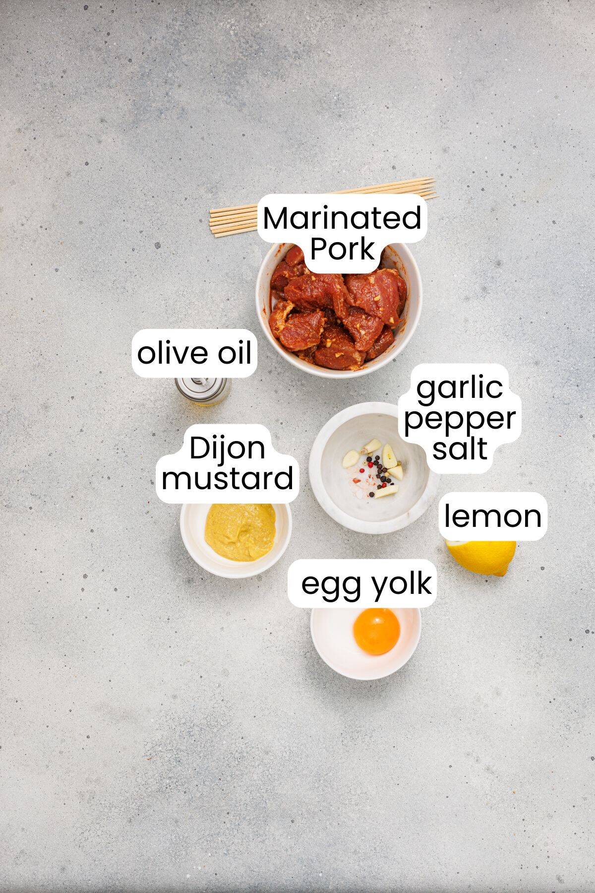 Top-down view of labeled ingredients for Grilled Pork Skewers: marinated pork, olive oil, Dijon mustard, egg yolk, garlic pepper salt, and lemon on a light gray surface—perfect for a homemade aioli.