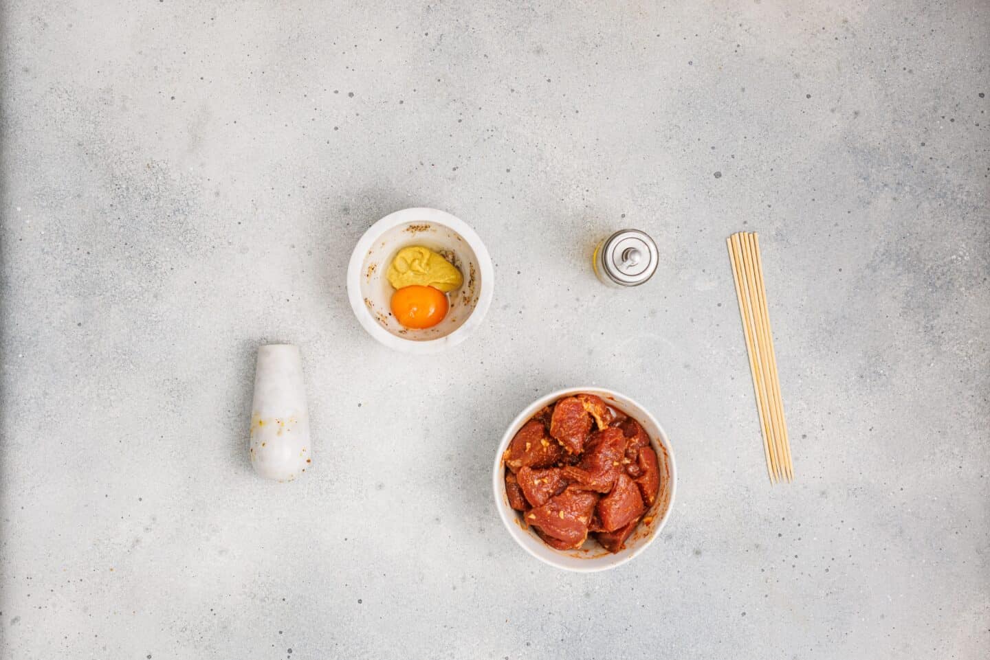 Raw marinated meat for Grilled Marinated Pork Skewers with Homemade Aioli sits in a bowl, beside an egg yolk with seasoning, mustard, and wooden skewers on a light countertop.