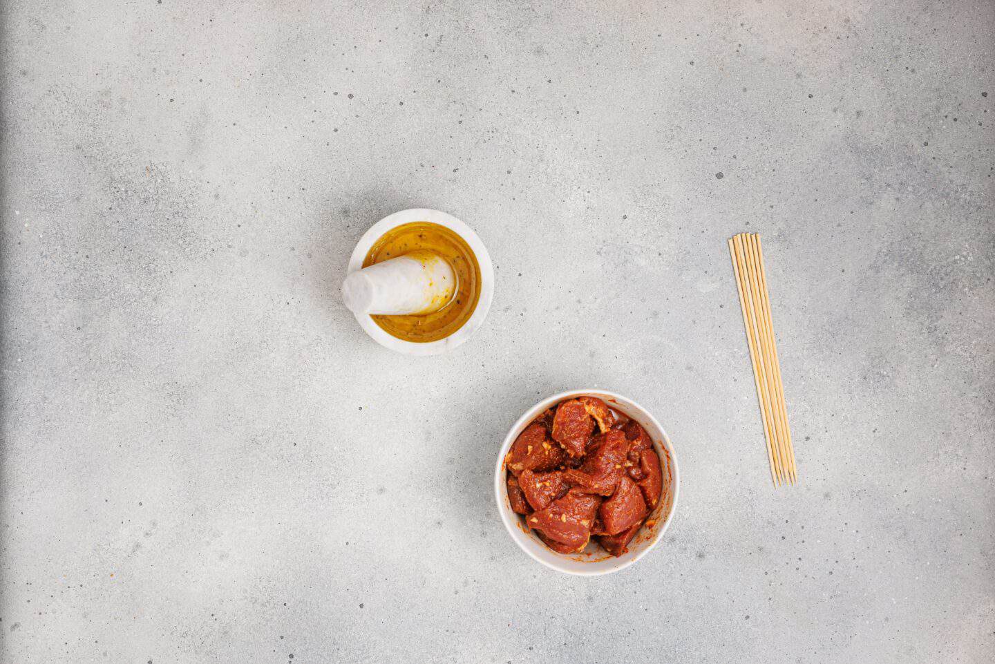 A bowl of Grilled Marinated Pork Skewers with Homemade Aioli, a mortar with pestle containing oil, and several wooden skewers rest on a light gray surface.