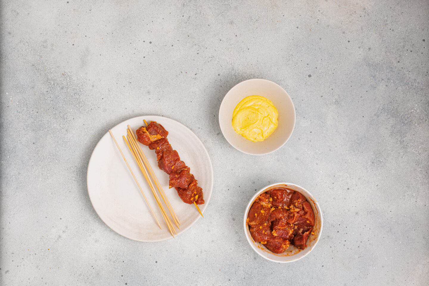 A plate with a raw Pork Skewer and several empty skewers, a bowl of Homemade Aioli, and a bowl of marinated meat on a light gray surface.