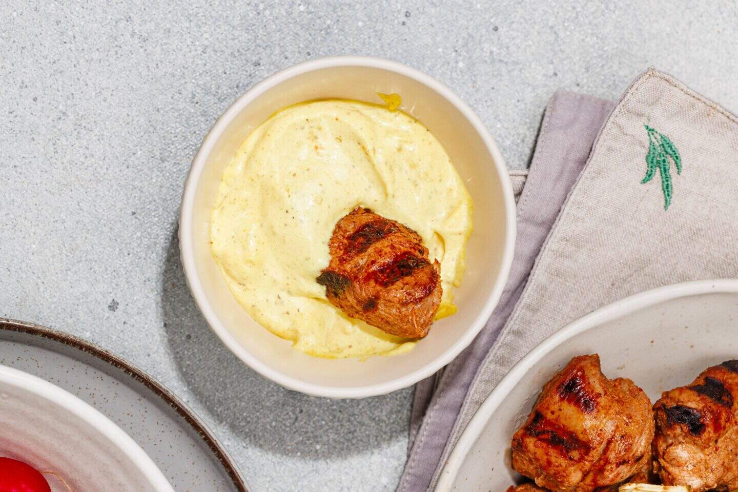 A bowl of creamy yellow homemade aioli with a piece of grilled marinated pork skewer partially dipped into it; a plate with more pork skewers is partially visible nearby.