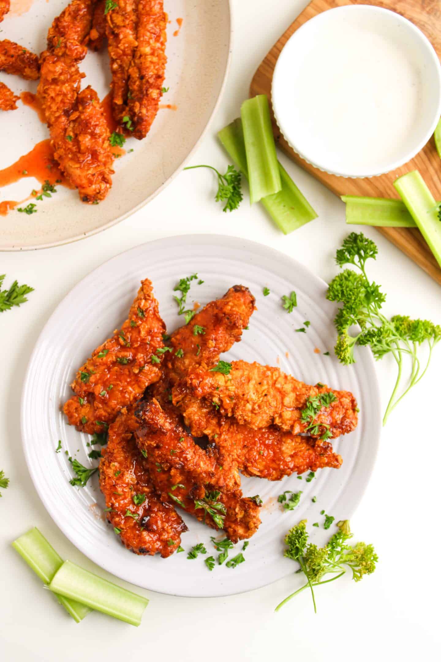 A plate of crispy hot honey chicken tenders garnished with parsley, served with celery sticks and a bowl of ranch dressing on the side.