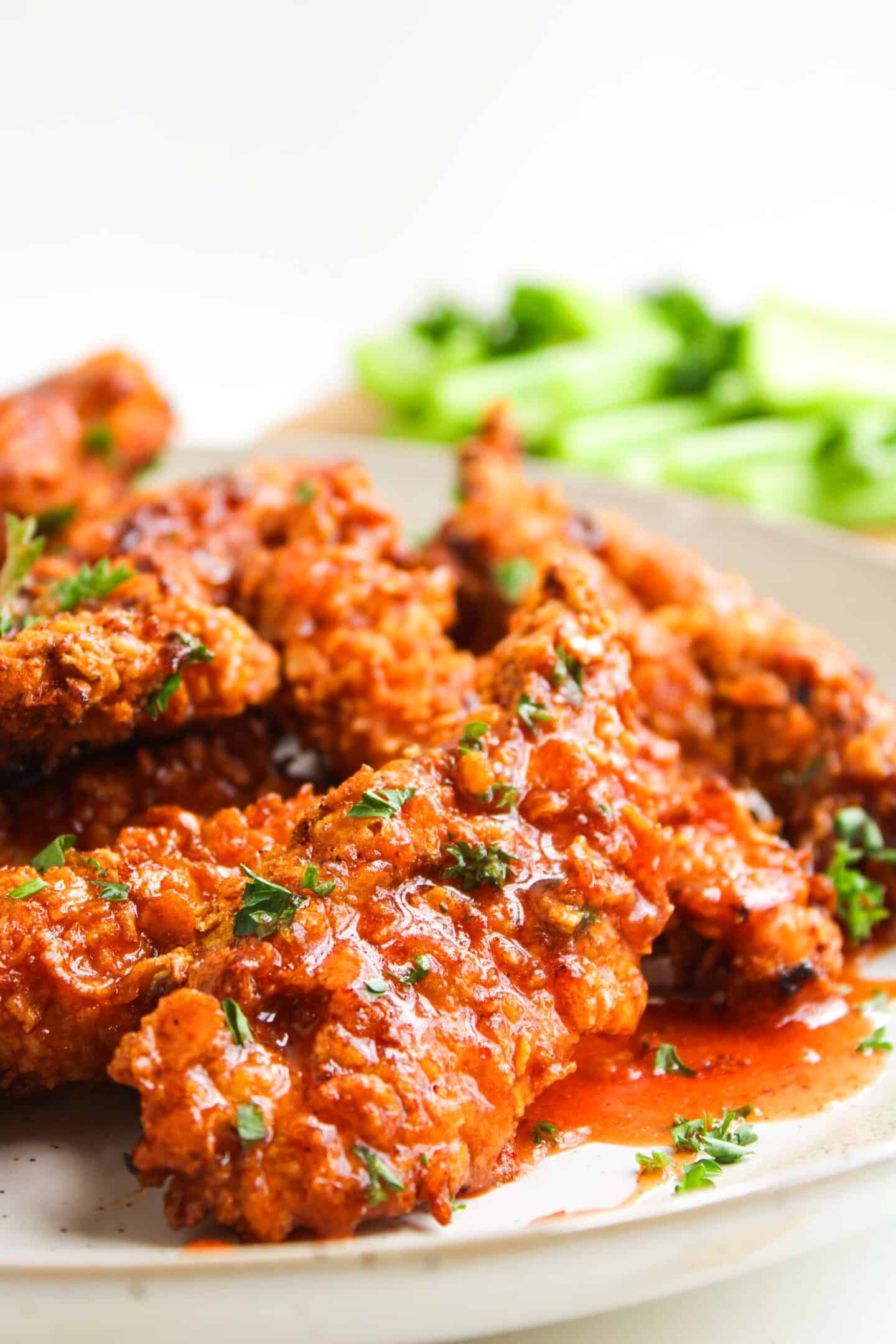 A plate of crispy air fryer chicken tenders coated in a hot honey red sauce, garnished with chopped herbs, with celery sticks in the background.