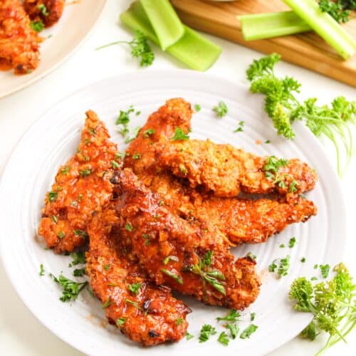 A white plate with several pieces of saucy Air Fryer Chicken Tenders, garnished with chopped parsley, surrounded by celery sticks and a bowl of hot honey dipping sauce.