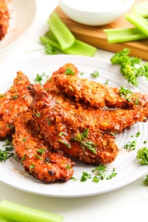 A plate of crispy Air Fryer Chicken tenders garnished with chopped parsley, served with celery sticks and a bowl of hot honey dipping sauce in the background.